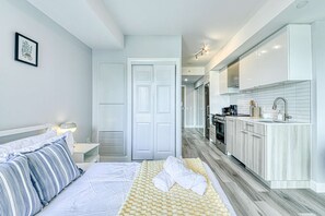 A modern bedroom with a bed, bedside table, and a small desk. There is a hallway leading to a kitchen with white cabinets and a sink. - RivetStays | The Jarvis Business Suite – Downtown Studio Suite (Toronto)