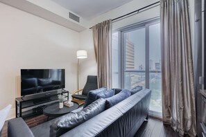 A modern living room with a large window, a flat-screen TV, a sofa, and a coffee table. - RivetStays | The Atmos Deluxe 2 Suite – 1 Bedroom Suite (Toronto)