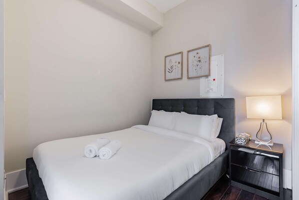 A neatly made bed with white linens and rolled towels, a dark upholstered headboard, a bedside table with a lamp, and two framed floral artworks on the wall. - RivetStays | The Atmos Deluxe 2 Suite – 1 Bedroom Suite (Toronto)