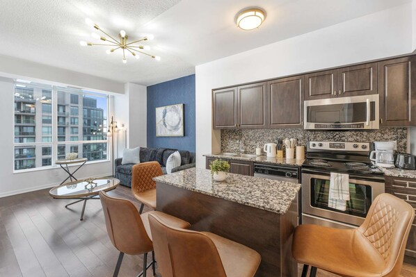 A modern kitchen with dark wood cabinets, stainless steel appliances, and a marble countertop. - RivetStays | The Neptune Signature Suite – 1 Bedroom + Den Condo (Toronto)