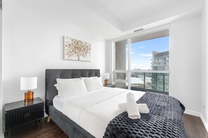 A modern hotel room with a large bed, a view of the city, and a cityscape visible through the window. - RivetStays | The Neptune Signature Suite – 1 Bedroom + Den Condo (Toronto)