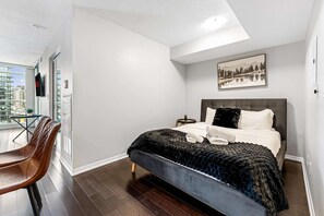 A modern bedroom with a large bed, a small bedside table, a chair, and a window with a city view. - RivetStays | The Neptune Design Suite – 1 Bedroom + Den Condo (Toronto)