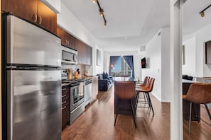 A modern kitchen with stainless steel appliances, a dining area with bar stools, and a view of the cityscape through large windows. - RivetStays | The Neptune City Suite – 1 Bedroom + Den Condo (Toronto)