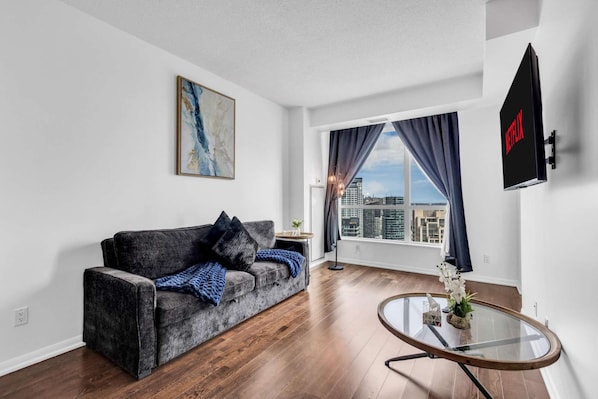 A modern living room with a grey sofa, a glass coffee table, and a city view through a large window. - RivetStays | The Neptune City Suite – 1 Bedroom + Den Condo (Toronto)