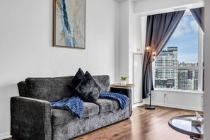 A modern living room with a grey sofa, a blue blanket, and a city view through large windows. - RivetStays | The Neptune City Suite – 1 Bedroom + Den Condo (Toronto)