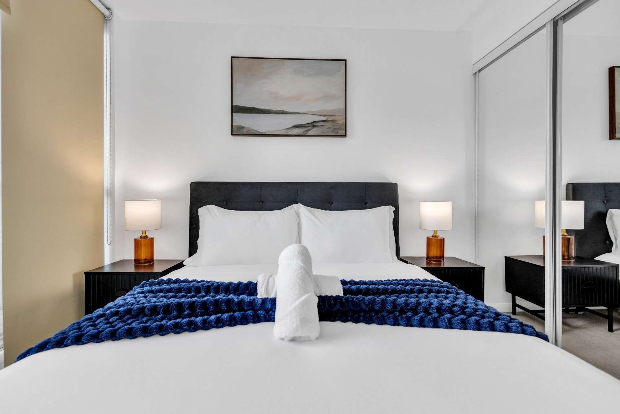 A hotel room with a large bed, two bedside tables with lamps, a wall-mounted painting, and a mirror.