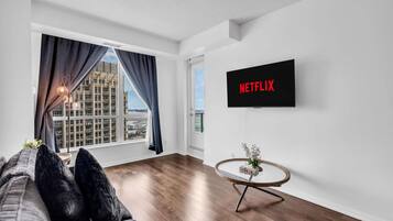 A modern living room with a large window, a flat-screen TV mounted on the wall, and a round coffee table.