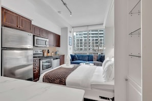 Ein kompaktes Hotelzimmer mit einer Küchenzeile, einem Bett mit weißer Bettwäsche, einem blauen Sofa und einem großen Fenster mit Blick auf die Stadt.