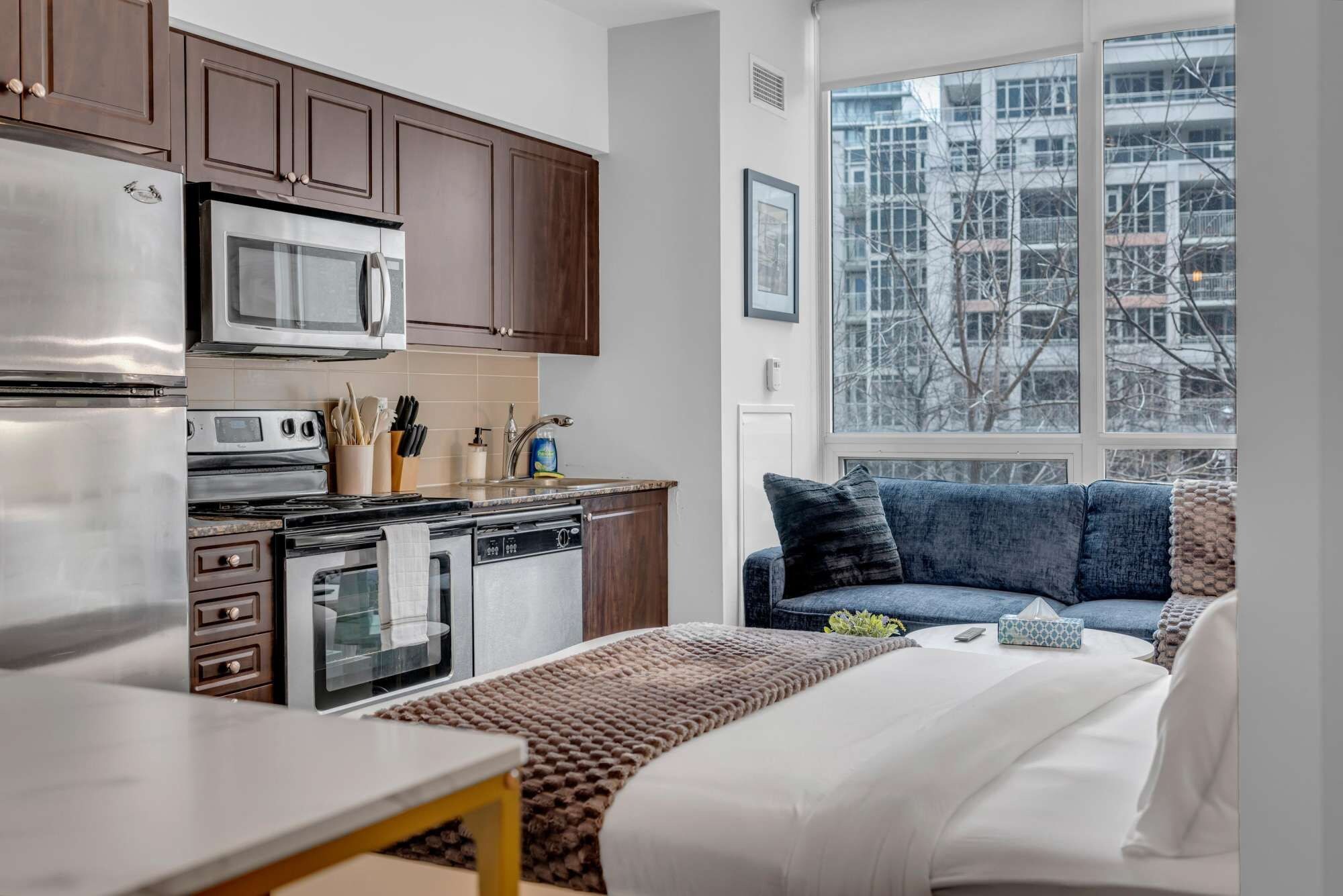 A compact kitchen with modern appliances, a dining area with a table, and a sofa with a view of the city through a large window.