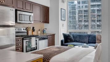 A compact kitchen with modern appliances, a dining area with a table, and a sofa with a view of the city through a large window.