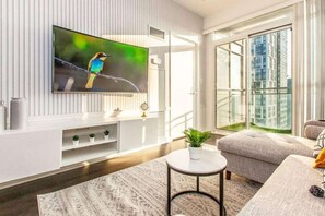 A modern living room with a flat-screen TV mounted on the wall, a grey sofa, a small round coffee table, and a large window offering a view of a cityscape. - RivetStays | The Parade 2 Executive Suite – 2 Bedroom Condo (Toronto)