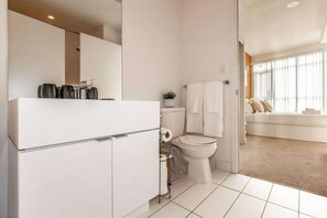 A modern bathroom with a toilet, sink, and a mirror. There is a window with blinds and a view of a living area with a sofa. - RivetStays | The Parade 2 Executive Suite – 2 Bedroom Condo (Toronto)