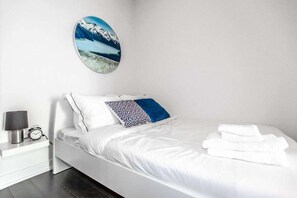 A modern bedroom with a bed, white bedding, a decorative wall piece, a bedside table with a lamp, and a clock. - RivetStays | The Parade 2 Executive Suite – 2 Bedroom Condo (Toronto)