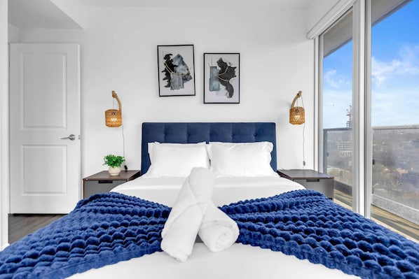 Une chambre d’hôtel avec un grand lit, deux œuvres d’art encadrées, une vue sur la ville par une grande fenêtre et un balcon.