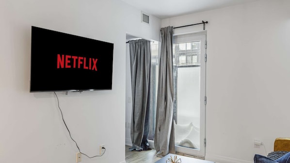 A room with a television mounted on the wall displaying 'NETFLIX' in red letters, a sliding glass door with grey curtains, and a wooden headboard.