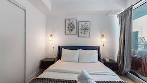 A bedroom with a bed, two bedside lamps, a window with curtains, and two framed floral artworks on the wall.
