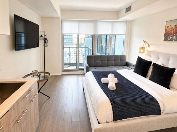 Une chambre d’hôtel moderne équipée d’un lit, d’une télévision, d’un bureau et d’une grande fenêtre donnant sur la ville.