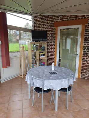 A covered patio with a round table and chairs, a television mounted on a brick wall, and a wooden cabinet with various items. - Cottage 'Le Gîte Du Bois Du Fil' 1km from GR210 (Vassonville)