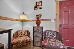 Ein Zimmer mit zwei Korbstühlen mit floralem Bezug, einem Holztisch, einer Lampe, einer Vase mit Blumen, einem Holzschrank und einer roten Tür.
