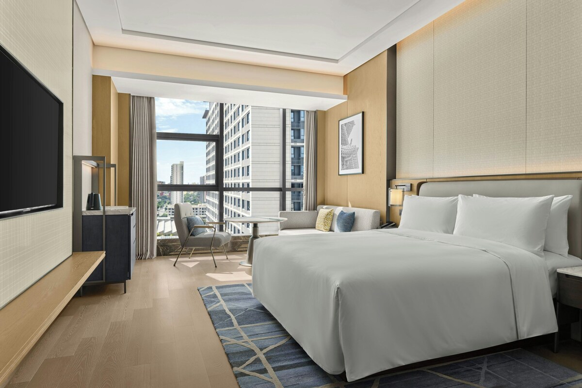 Habitación de hotel con una cama grande, un escritorio, una silla, un televisor y vistas a la ciudad.