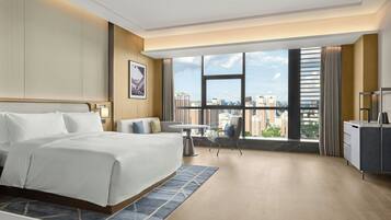 A hotel room with a large bed, a desk, a chair, and a view of the city through large windows.