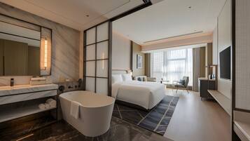 Una habitación de hotel moderna con una cama grande, un baño separado con bañera, una zona de estar y un televisor.