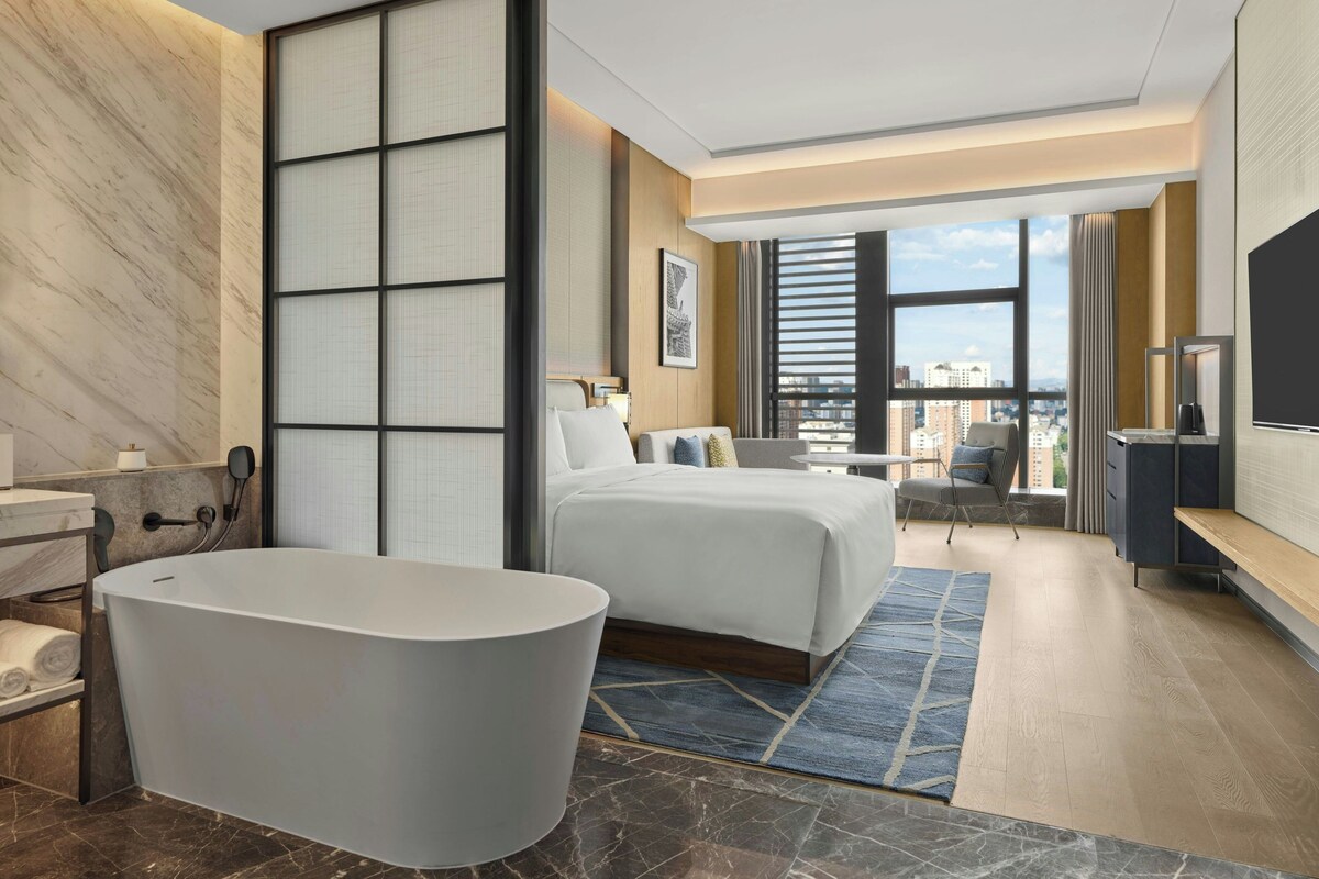 Habitación de hotel moderna con una cama grande, una bañera, vistas a la ciudad y un televisor de pantalla plana.