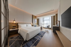 A modern hotel room with a large bed, bedside tables, a desk, a chair, a TV, and a window with curtains.