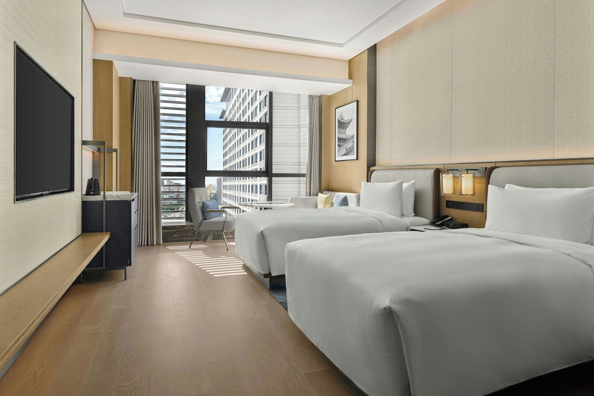 Habitación de hotel con dos camas, televisión, escritorio, silla y vistas a edificios.