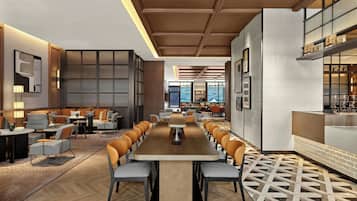 Un hall d’hôtel moderne avec une longue table commune, des espaces de détente et une vue sur le paysage urbain grâce à de grandes fenêtres.