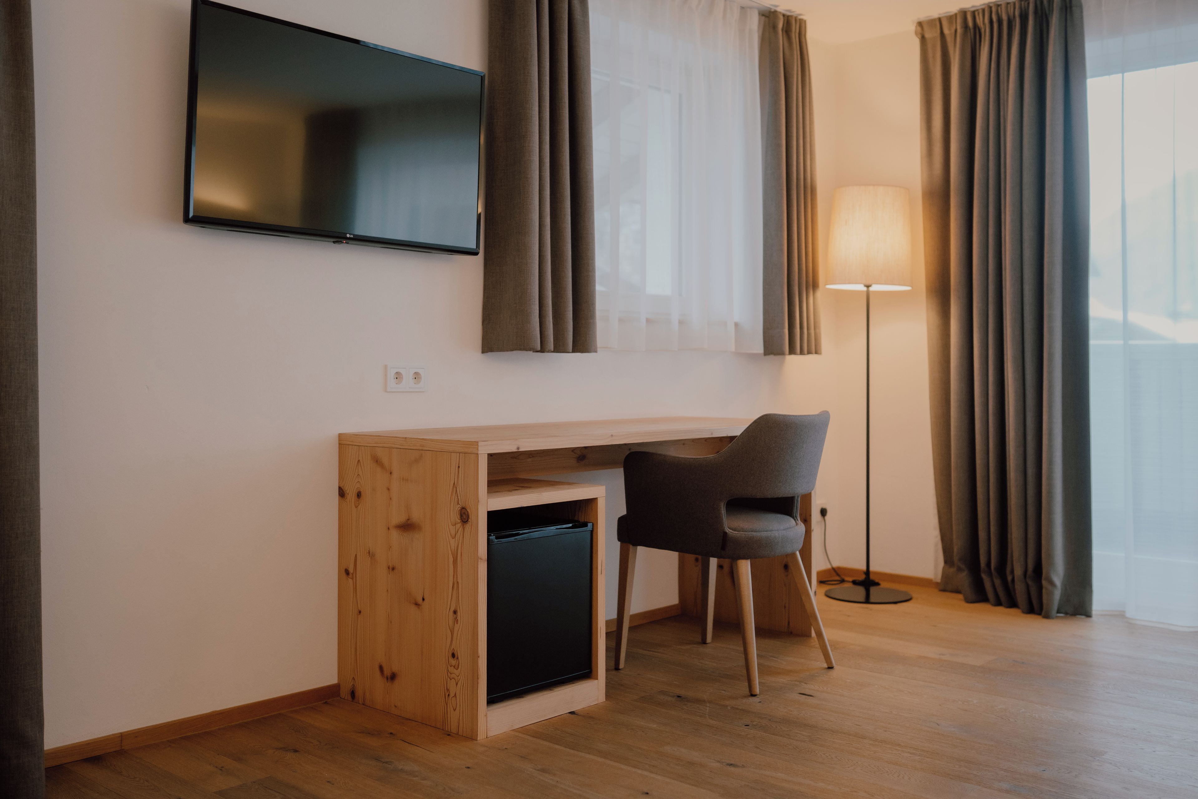 Una habitación moderna con un escritorio de madera, una silla, un televisor montado en la pared y una lámpara de pie.
