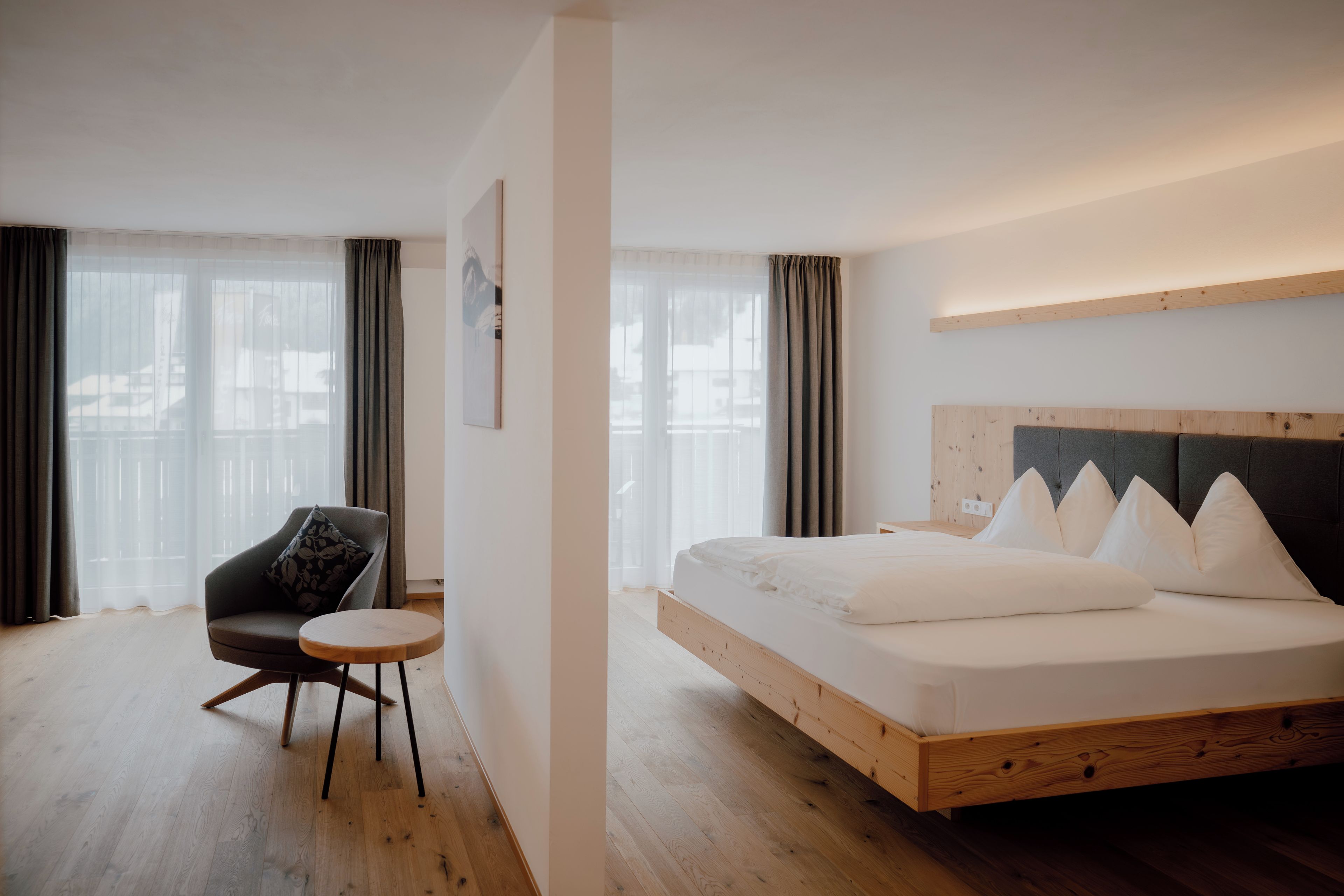 A hotel room with a bed, a chair, a small table, and large windows with curtains.