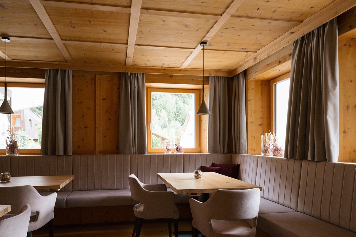 A cozy cabin interior with a wooden ceiling, a dining table with chairs, a bench, and large windows with curtains.