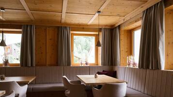 A cozy cabin interior with a wooden ceiling, a dining table with chairs, a bench, and large windows with curtains.
