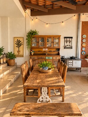 Un comedor con una larga mesa de madera, sillas y un banco. Hay un armario con puertas de cristal y un tendedero con utensilios de cocina.