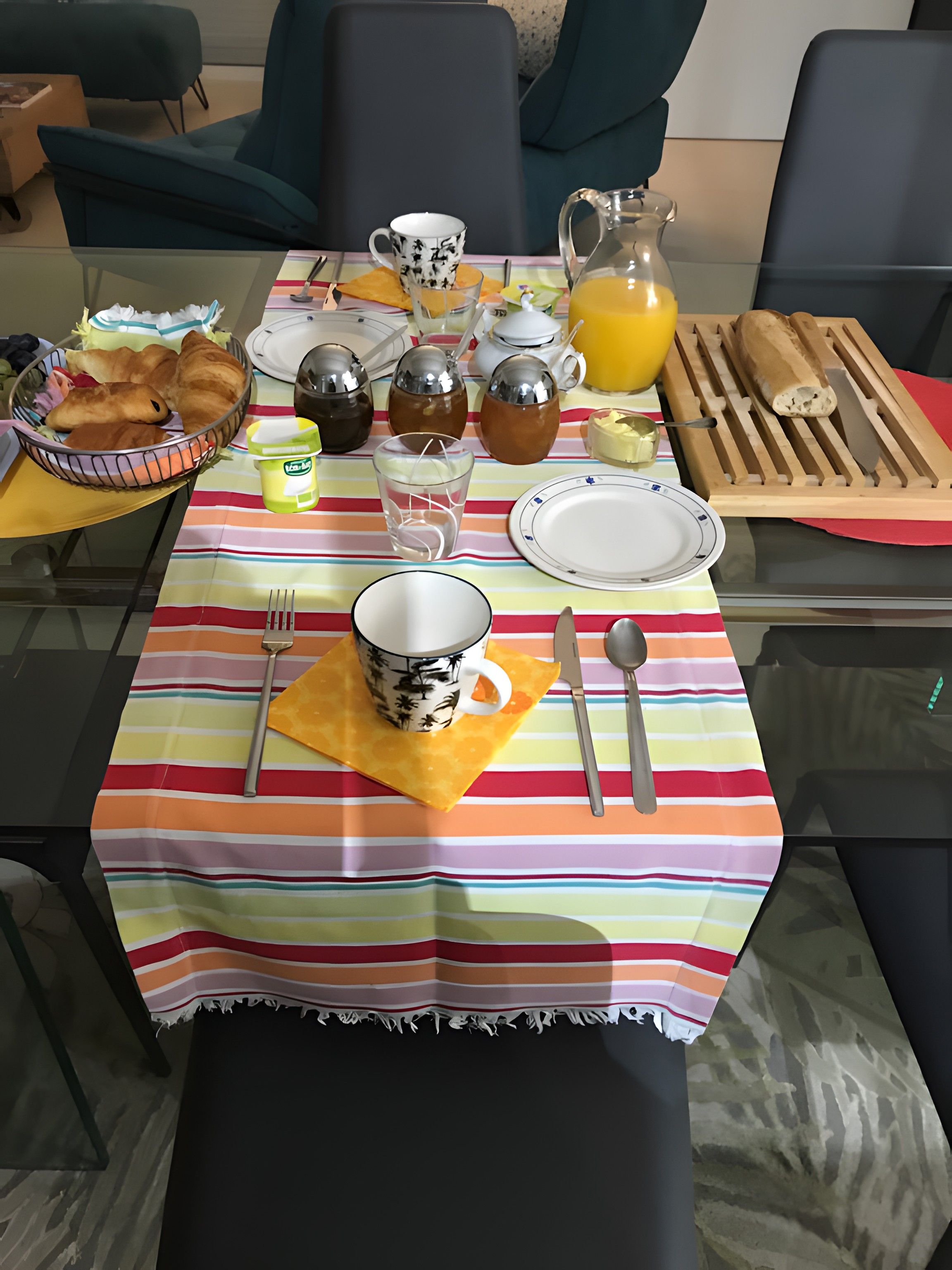Une table de petit-déjeuner dressée avec des assiettes, des couverts, un pichet de jus d’orange, un panier de croissants et une théière.