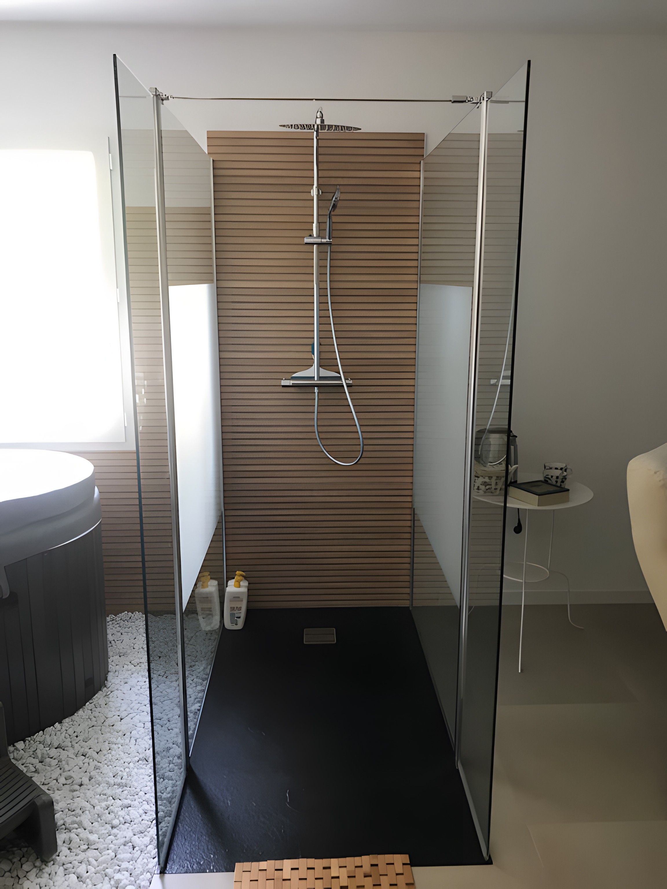 Une salle de bain moderne avec une douche à parois de verre, un mur recouvert de panneaux en bois, un lavabo rond et une petite table sur laquelle se trouvent une tasse et un livre.