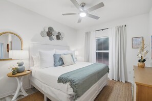 Ein Schlafzimmer mit Bett, Nachttisch, Spiegel, Deckenventilator, Fenster mit Vorhängen und Wanddeko.