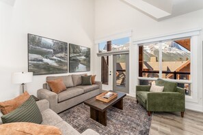 A living room with a sofa, armchairs, a coffee table, and a large window with a mountain view. - Mountain Eye Candy | Penthouse with Pool and Hot Tub (Canmore)