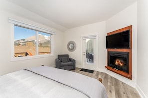 A bedroom with a bed, a fireplace, a television, and a view of mountains through a window. - Mountain Eye Candy | Penthouse with Pool and Hot Tub (Canmore)