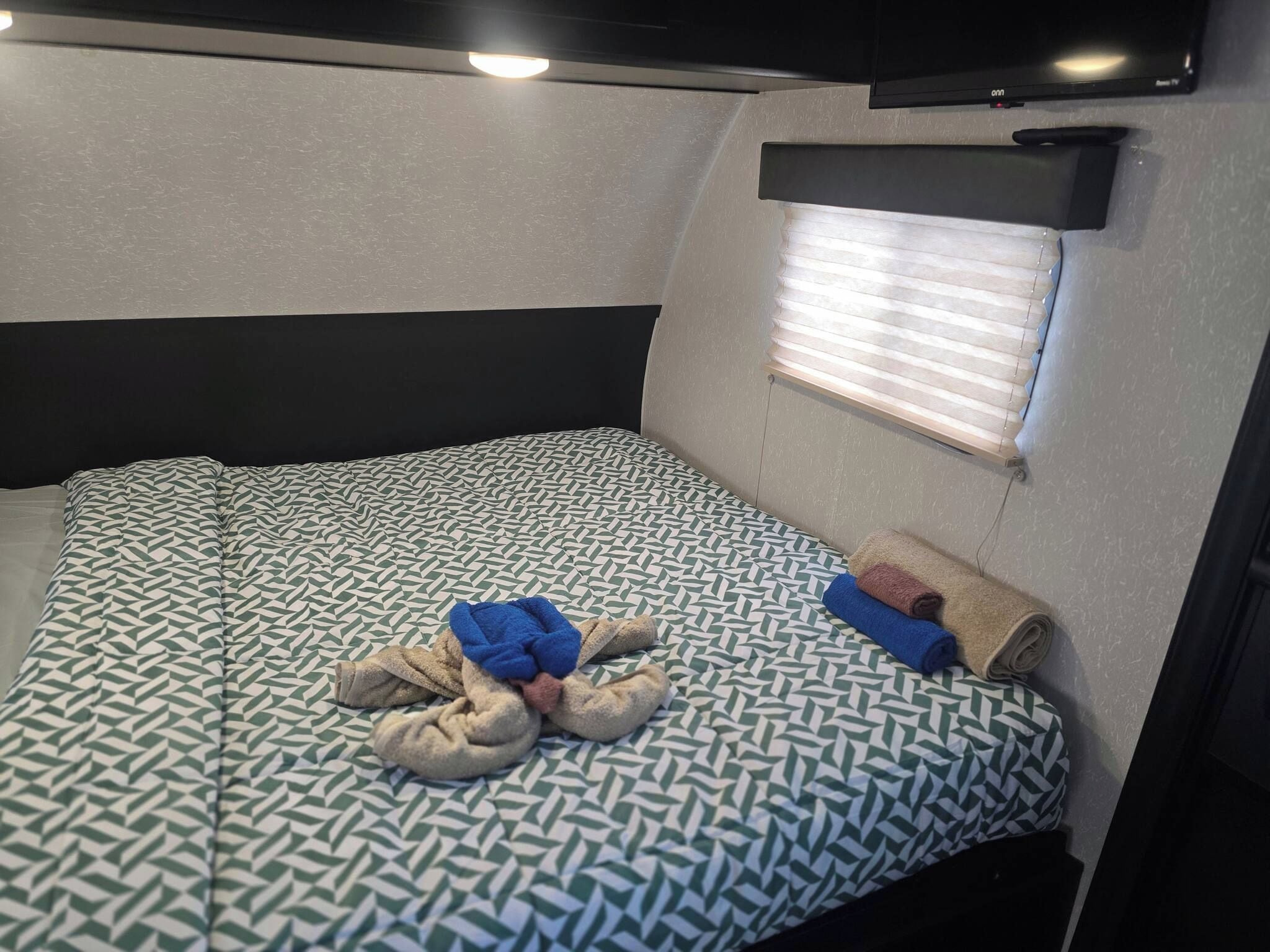 A compact bed with a patterned bedspread, a window with blinds, and a television mounted above the bed.
