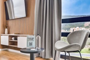 A modern hotel room with a wooden panel wall, a grey upholstered chair, a small round table with a water bottle and a brochure, and a large window with a city view. - w* | Dreamy & Cozy Loft in the Heart of Bogotá (Bogotá)