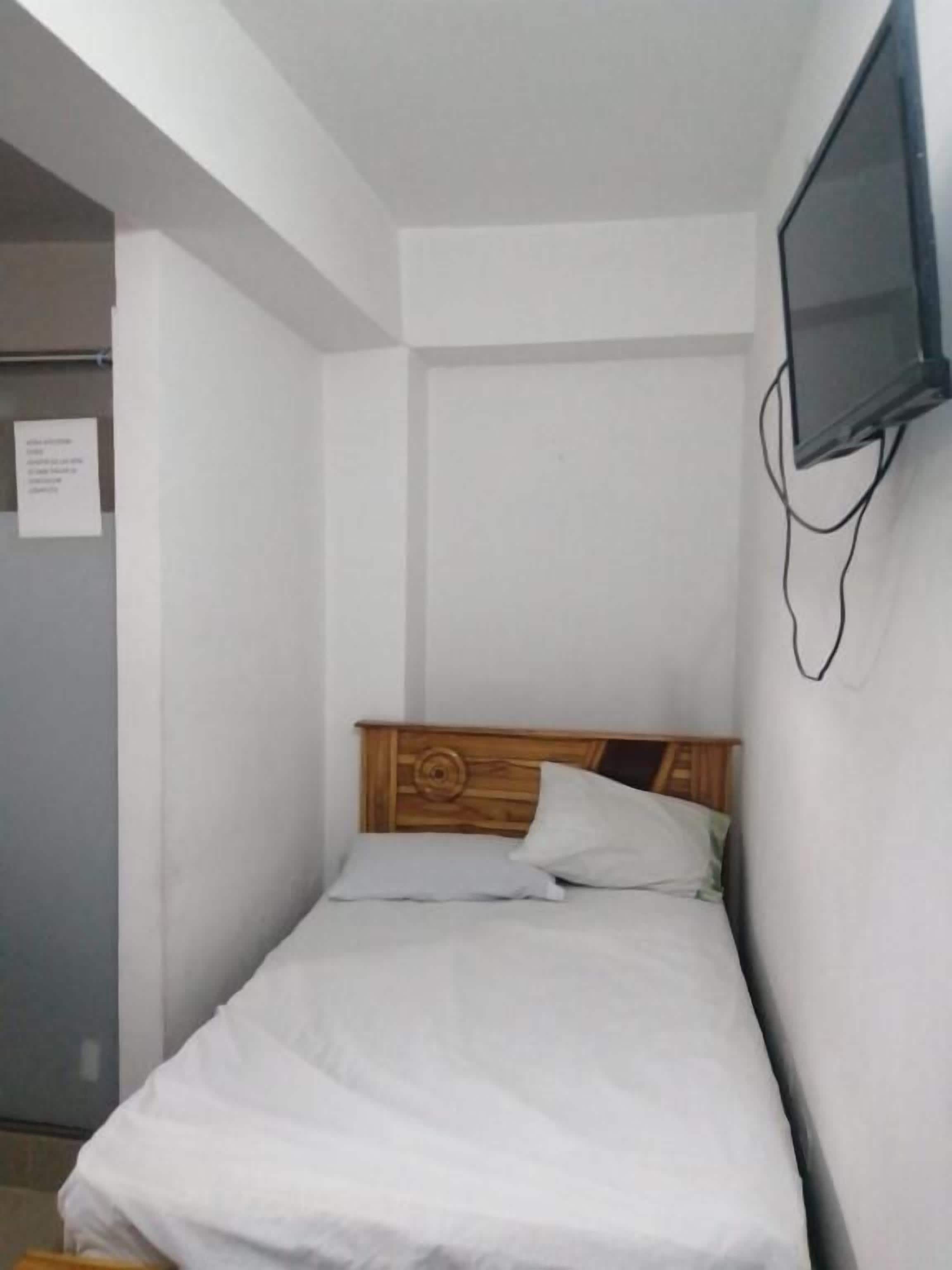 A small, single-bed room with a wooden headboard, a flat-screen TV mounted on the wall, and a door with a sign.
