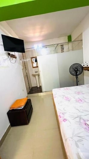 Een slaapkamer met een bed, een ventilator, een klein tafeltje en een opbergkist.