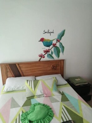 A bed with a geometric patterned bedspread, a green ruffled pillow, and a wall decoration of a colorful bird on a branch.