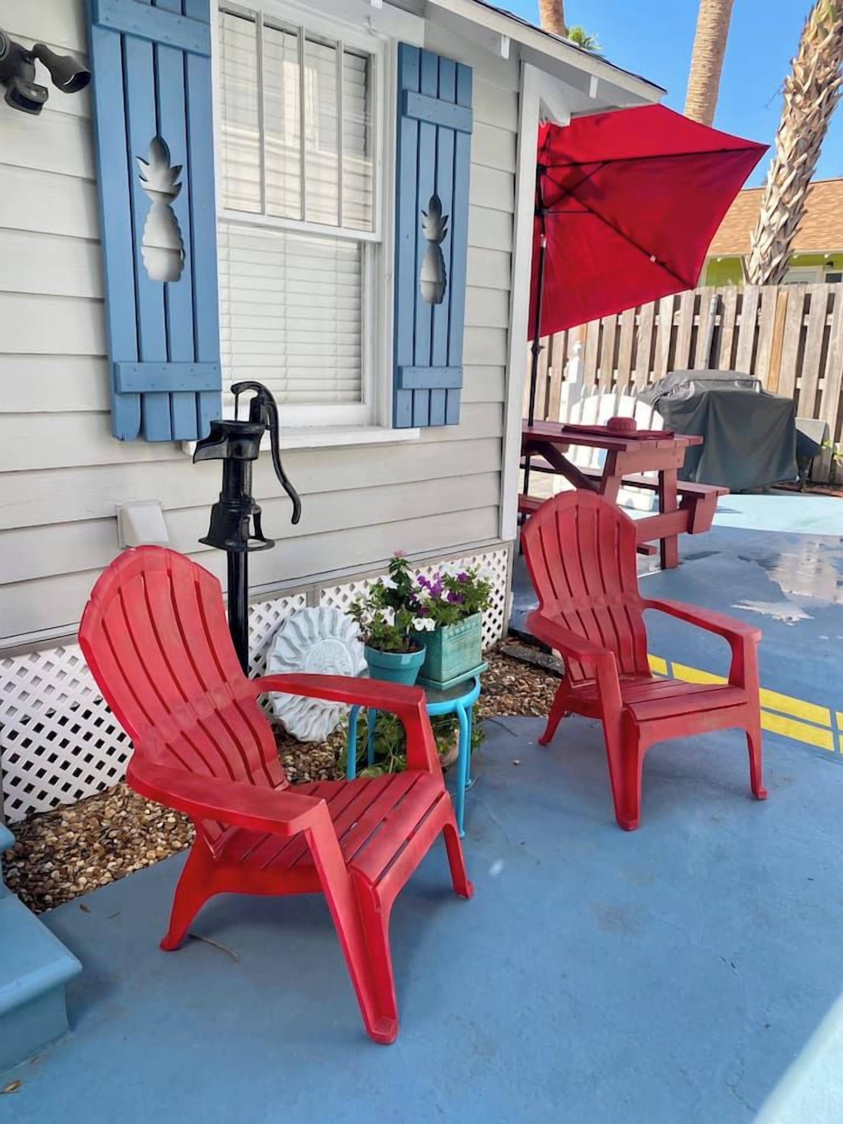Two red Adirondack chairs, a small table, and a red umbrella outside a house with blue shutters.