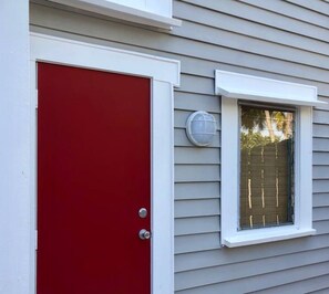 Une porte rouge avec un cadre blanc, une fenêtre avec des stores et un petit luminaire fixé au mur.