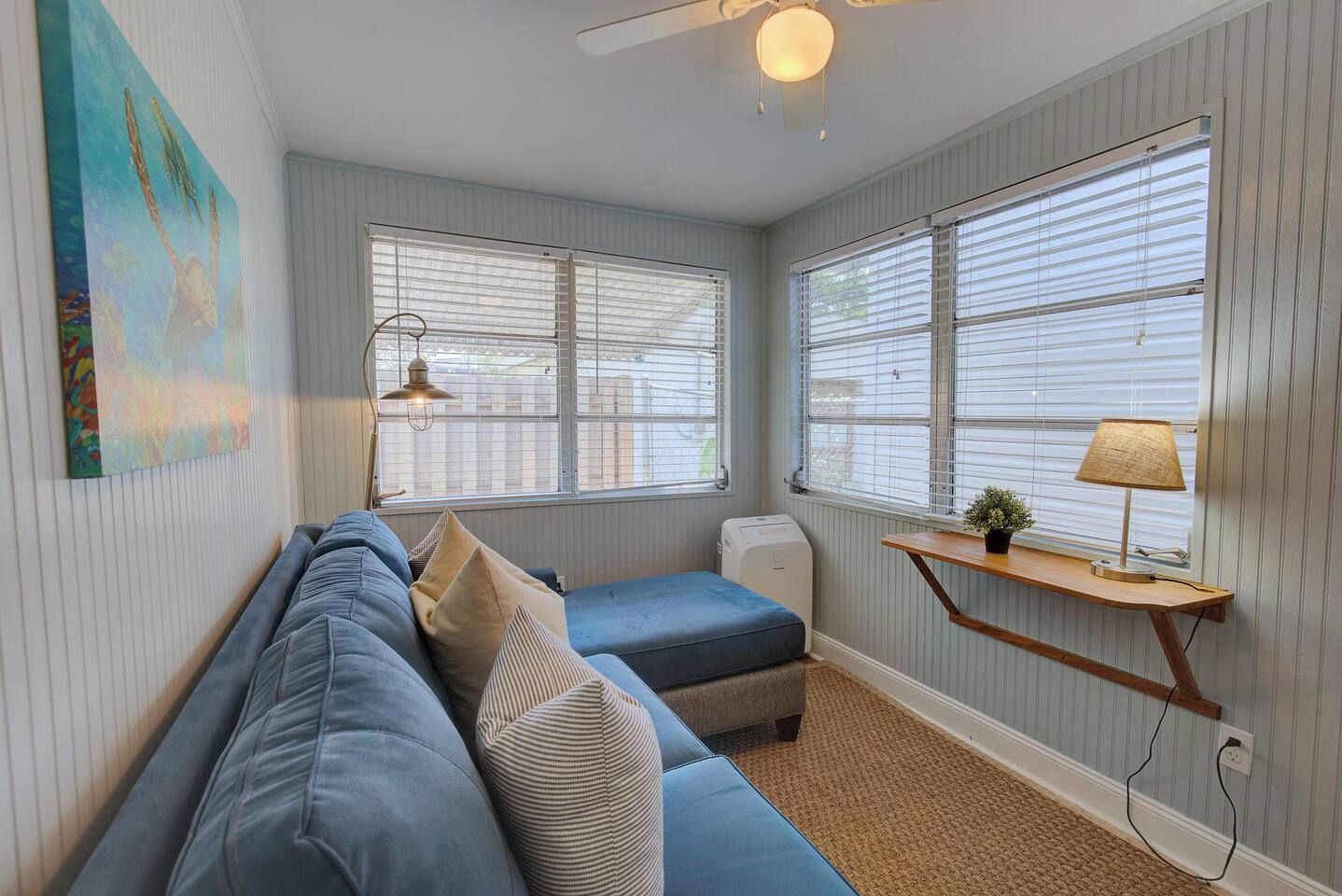 A compact room with a blue sofa, a small ottoman, a lamp on a wooden shelf, and a ceiling fan.