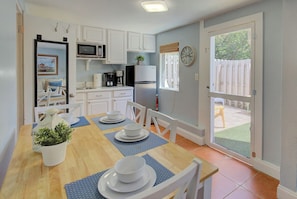 A compact kitchen with white cabinets, a microwave, a coffee maker, and a refrigerator. There is a dining table with blue placemats and white plates. A clock is on the wall, and a door leads to a screened outdoor area. - Downtown Dunedin 917-1/2 (Dunedin)