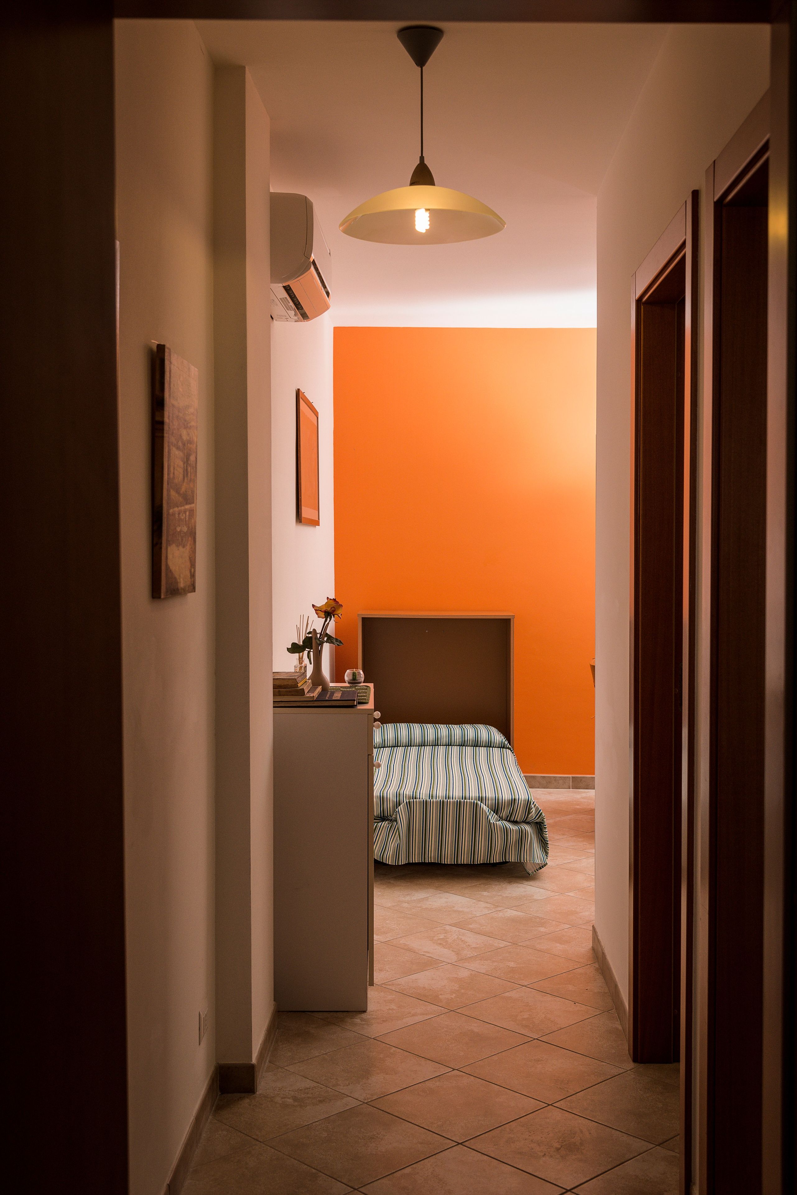 Un couloir étroit mène à une chambre dont un des murs est orange, avec un lit recouvert d’une couette à rayures, une table de chevet ornée d’un vase et un luminaire suspendu au plafond.